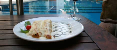 Una mesa de comedor con un plato de comida, una copa de vino tinto y una piscina al fondo.