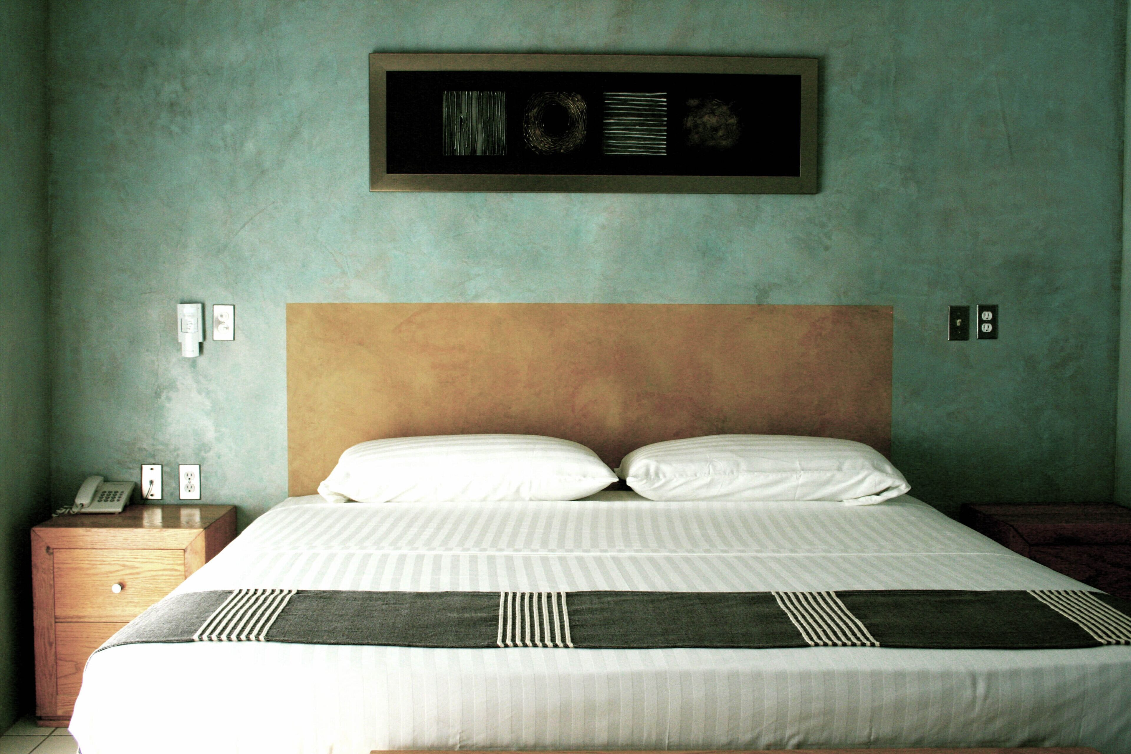 A neatly made bed with a headboard, a bedside table with a phone and a small clock, and a framed picture above the bed.