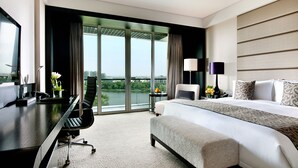 A hotel room with a large bed, a desk, a chair, and a view of a cityscape through large windows.