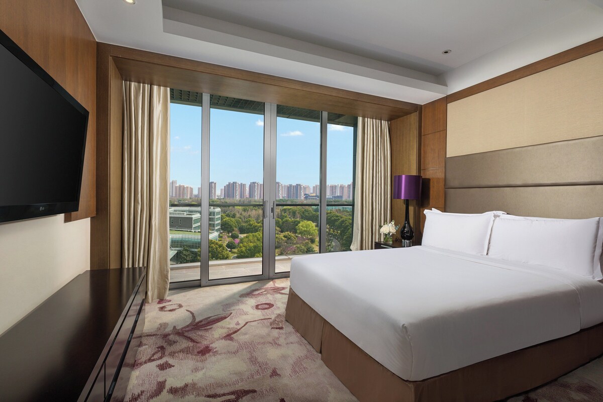 Habitación de hotel con una cama grande, televisor de pantalla plana, vista del paisaje urbano a través de ventanales del suelo al techo y un diseño moderno.