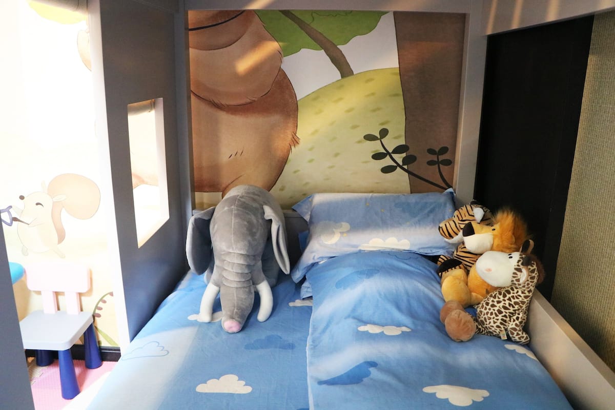 Habitació infantil amb un llit, animals de peluix i un mural d'un arbre i núvols.
