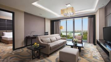 A modern hotel room with a sofa, a coffee table, a TV, and a large window offering a city view.