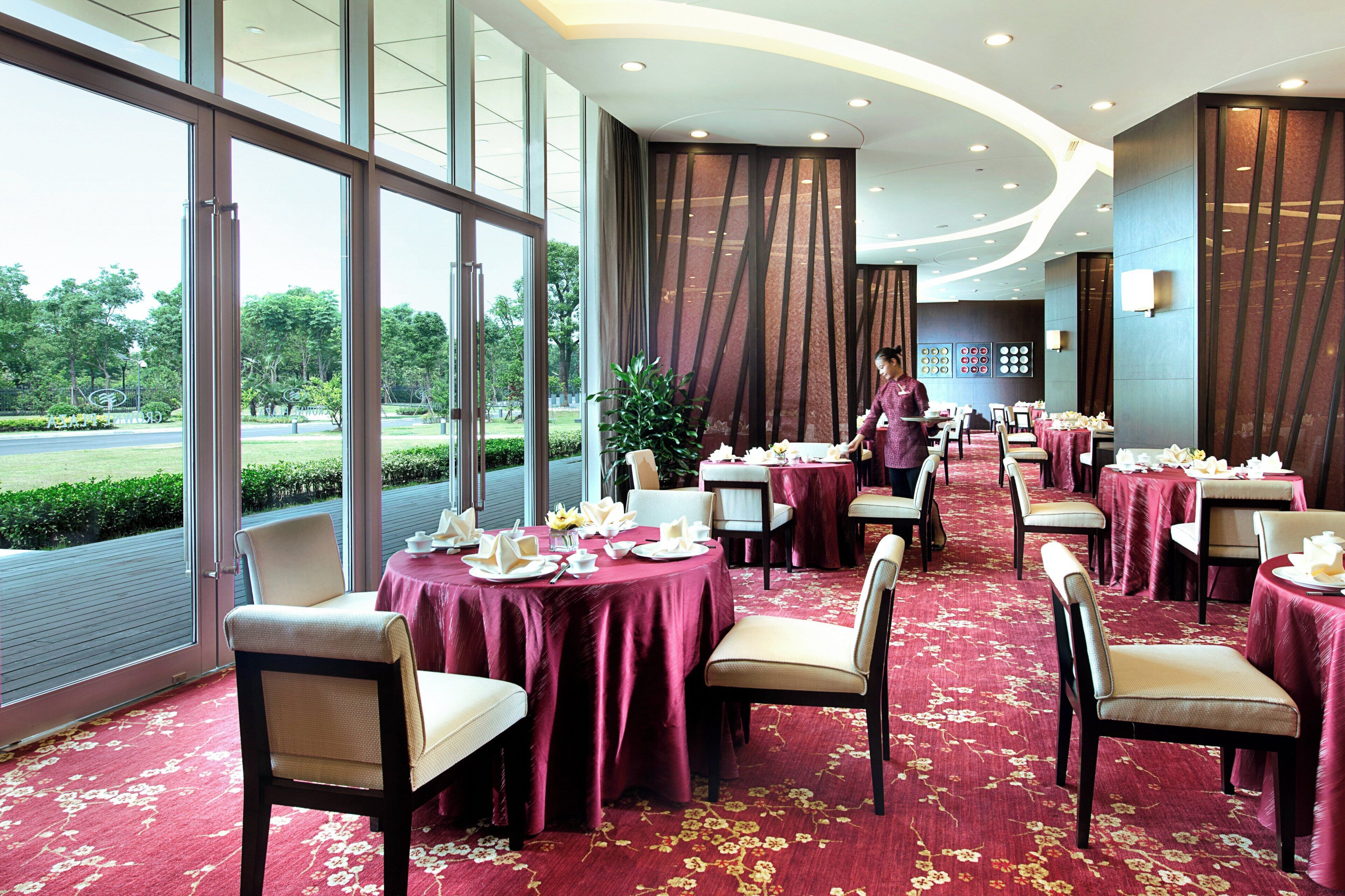 A restaurant with tables set for dining, large windows, and a view of greenery outside.