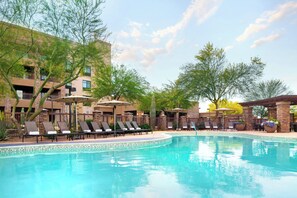 A pool area with lounge chairs and umbrellas, surrounded by a building with balconies and trees.