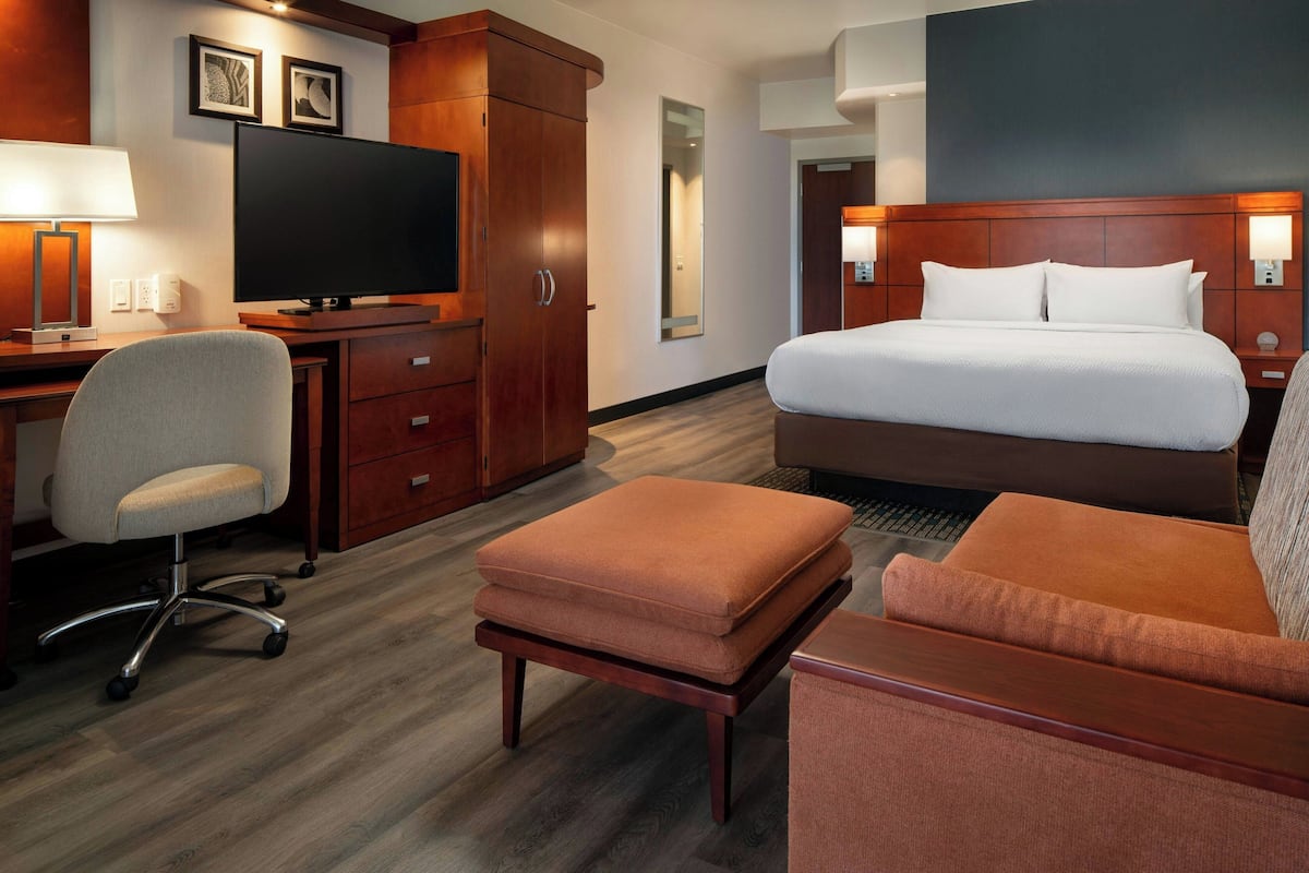 A hotel room with a bed, a desk with a TV, a chair, and an ottoman.