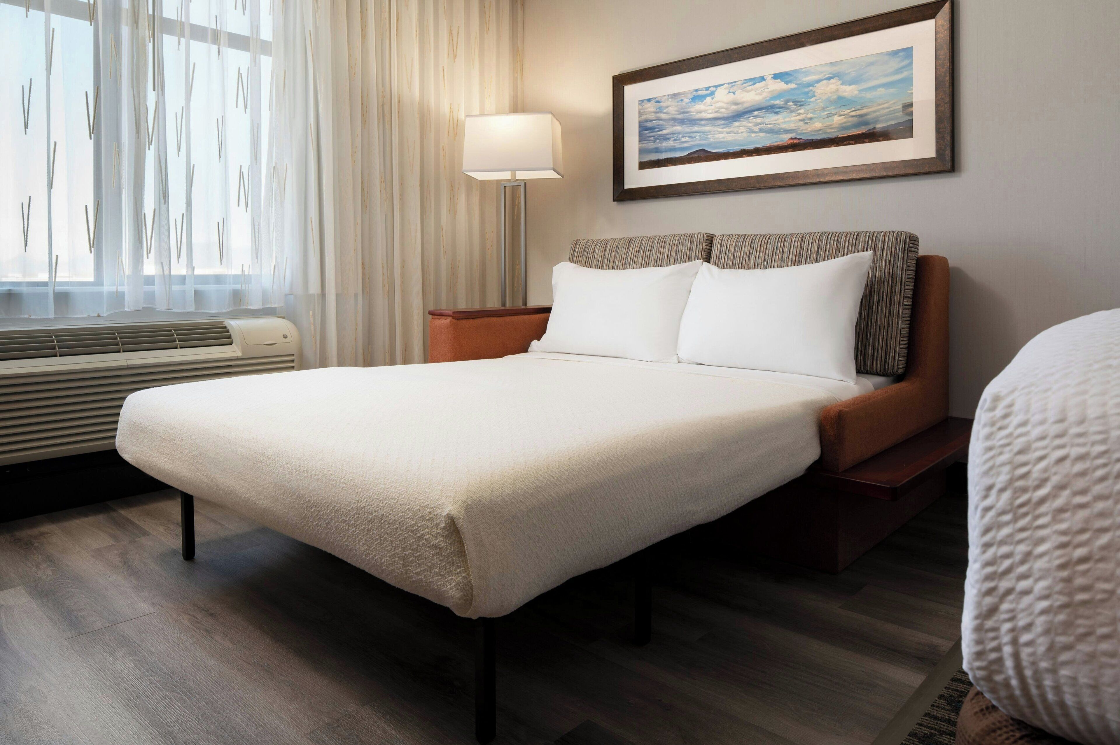 A neatly made bed with white linens, a bedside lamp, and a framed picture on the wall.