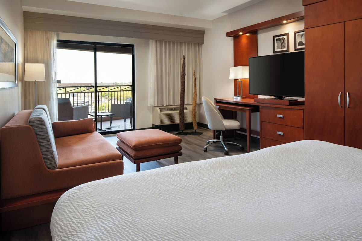 A hotel room with a large bed, a sofa, a desk, a television, and a balcony with outdoor seating.