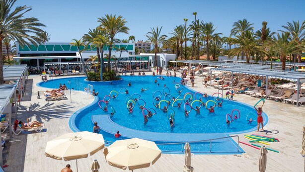 Zona de piscina con diversas actividades acuáticas, palmeras y sillones de descanso.