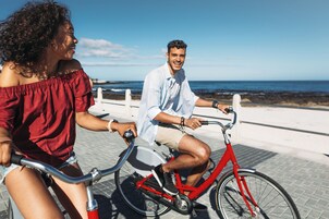 Two people riding a red bicycle on a paved path with a coastal view.