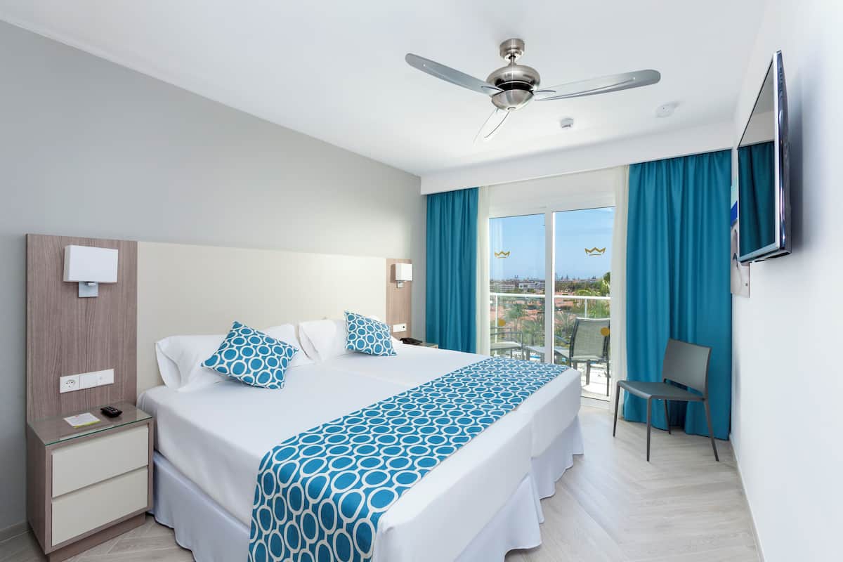 A hotel room with a bed, a nightstand, a chair, a TV, and a balcony with blue curtains.