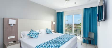 A hotel room with a bed, a nightstand, a chair, a TV, and a balcony with blue curtains.