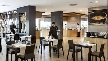 A modern restaurant with dark wooden tables and chairs, a central serving area, and a large wall-mounted menu display.