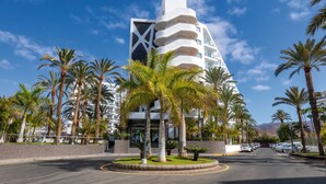 Un hotel moderno con un llamativo logotipo blanco y rojo, rodeado de palmeras y un cielo azul despejado.