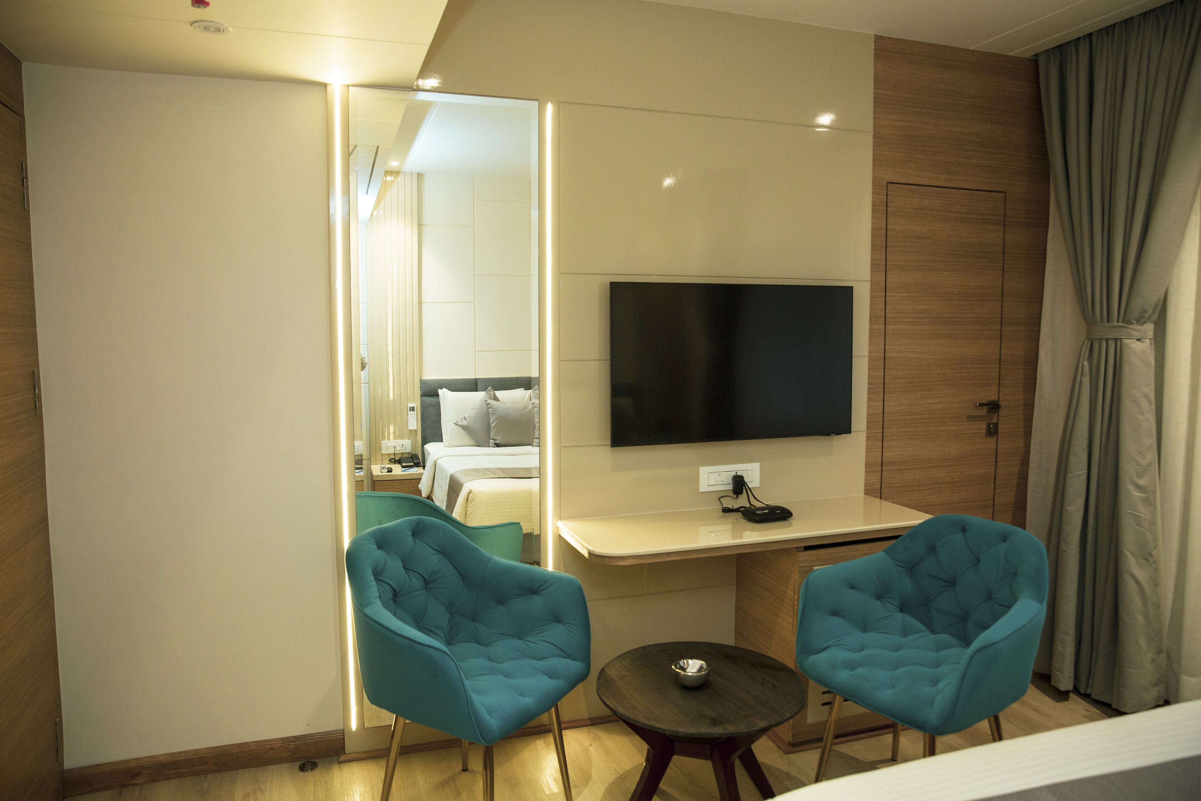 A modern hotel room with a large flat-screen TV, two matching blue armchairs, a small round table, and a wooden wardrobe.