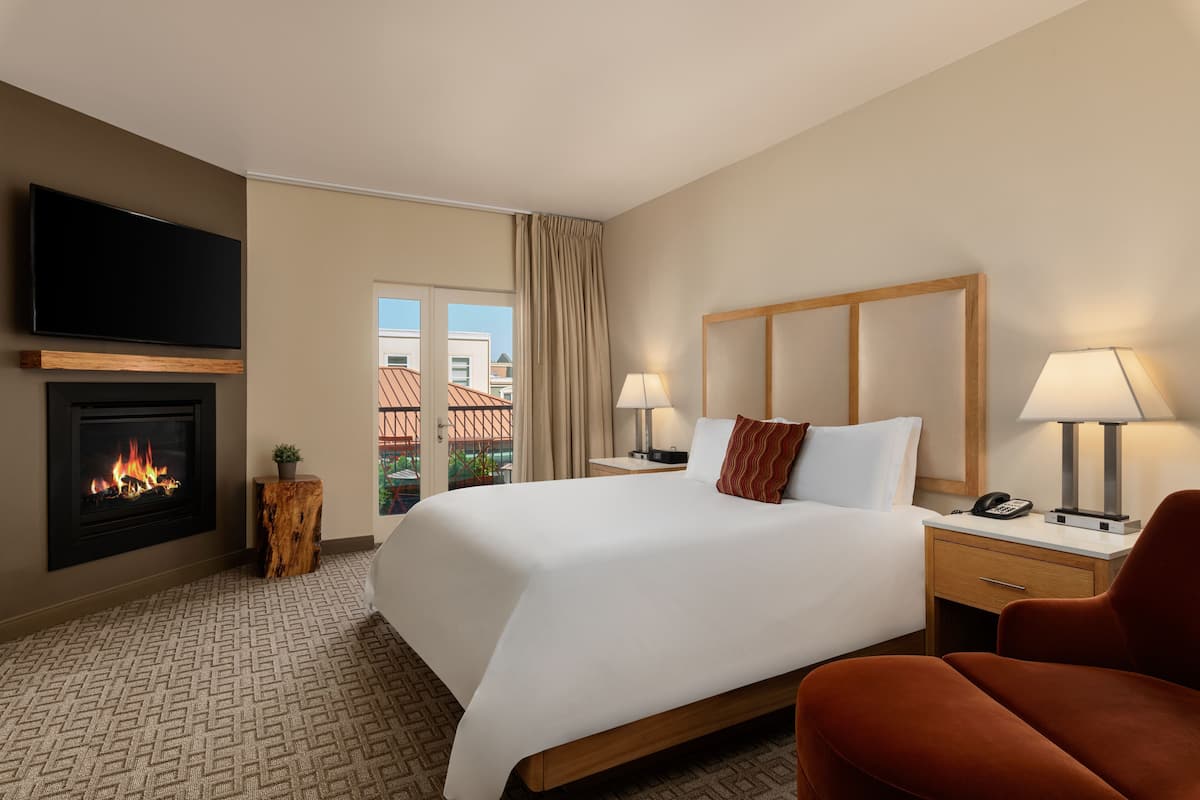 A hotel room with a bed, bedside lamps, a TV, a fireplace, and a view of buildings outside.