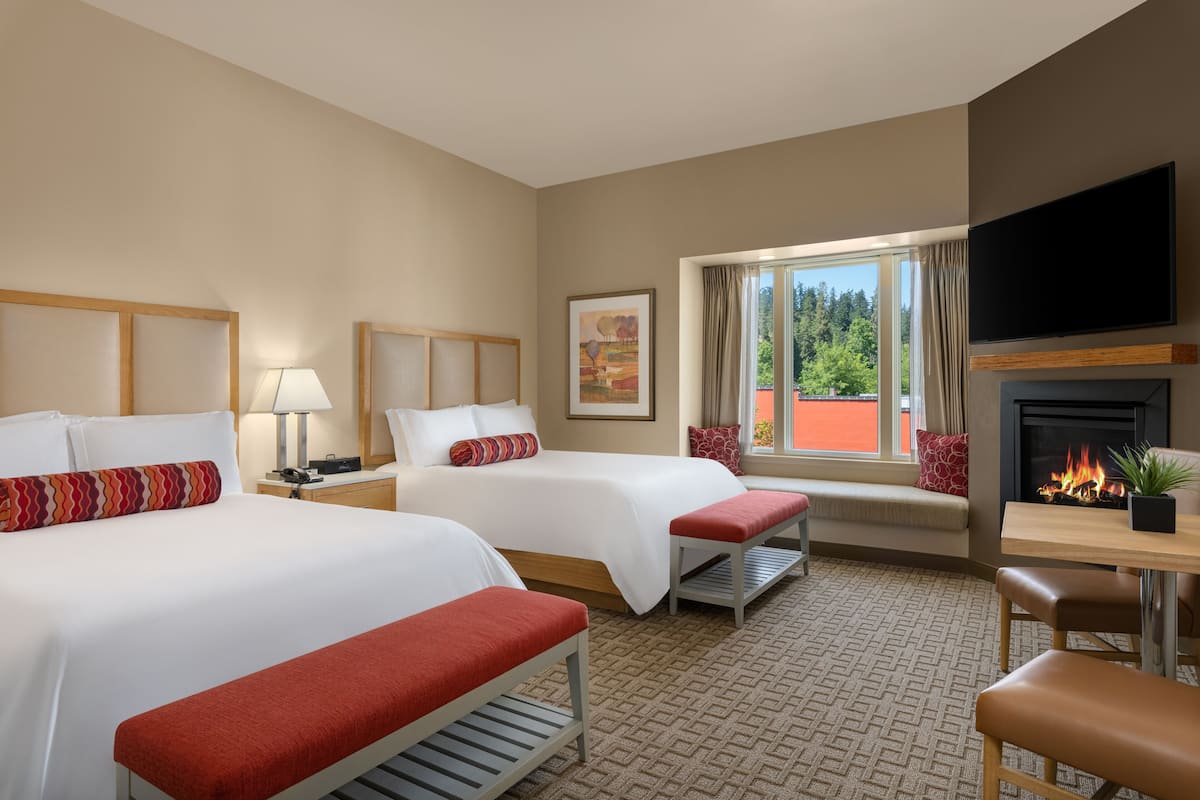 A hotel room with two beds, a TV, a fireplace, and a window with a view of trees.