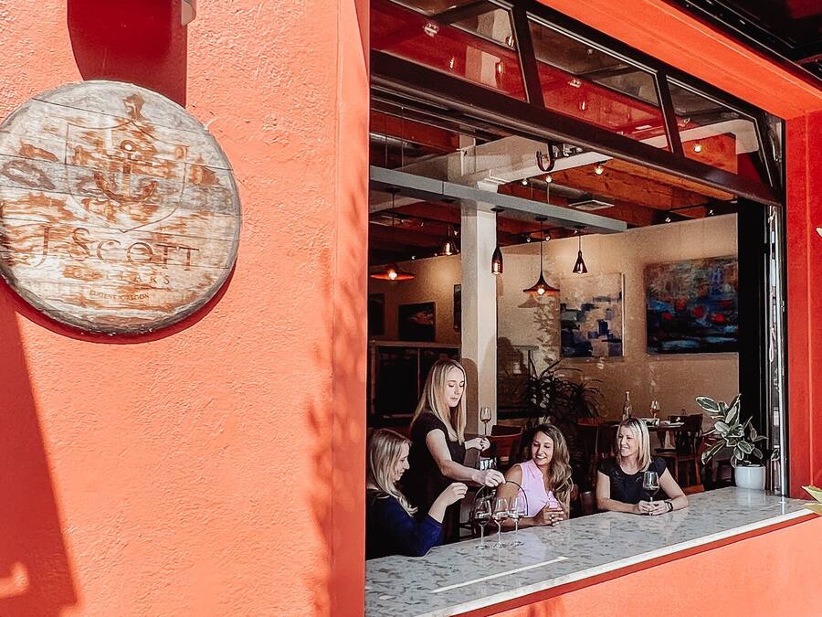An outdoor seating area with a red wall, a round wooden sign, and a view into the interior of a restaurant.