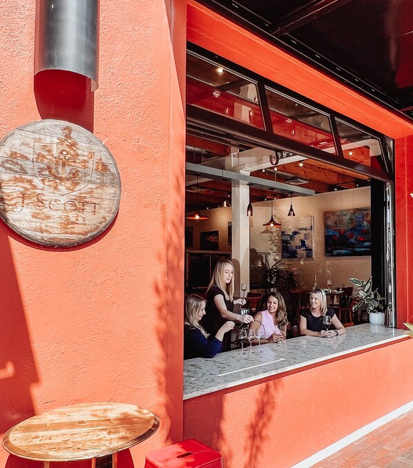 Un área de estar al aire libre con una pared roja, un letrero de madera redondo y vistas al interior de un restaurante.