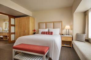 Habitación de hotel con una cama grande, un armario de madera, una mesita de noche con lámpara, un banco y vistas al baño.