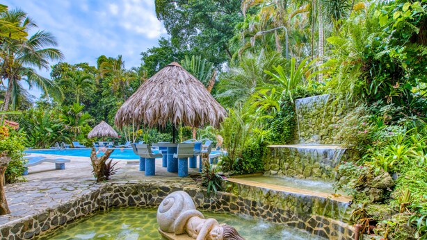 Un resort tropical con piscina, cabañas de techo de paja y una fuente de piedra con una escultura.