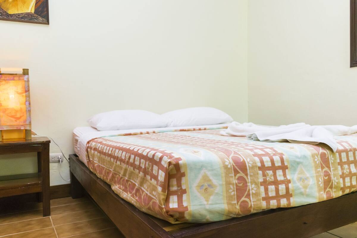 Een net opgemaakt bed met een beddekleed met patroon, een nachtkastje met een lamp, en een aan de muur hangend schilderij.