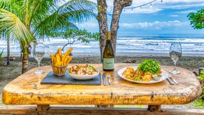 Una mesa de madera con dos platos de comida, una botella de vino y vasos, sobre una playa con palmeras y el océano al fondo.