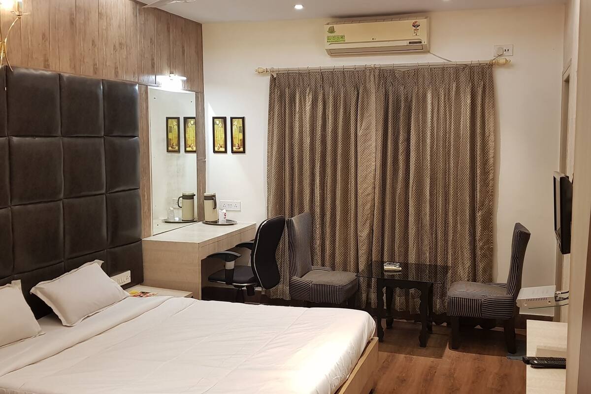 A hotel room with a large bed, a desk, two chairs, a TV, and an air conditioning unit.
