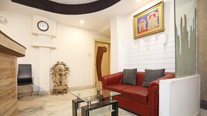 A living room with a glass coffee table, red sofa, and a statue. There is a clock on the wall and a framed picture above the sofa. - Hotel Rushabh Home (Kolkata)