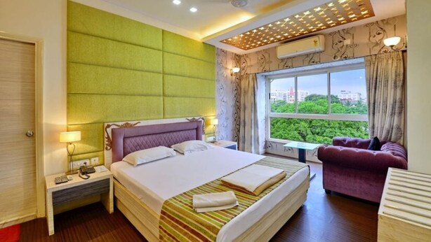 A hotel room with a large bed, a sofa, a window with a city view, a ceiling fan, and an air conditioning unit. - Hotel Rushabh Home (Kolkata)