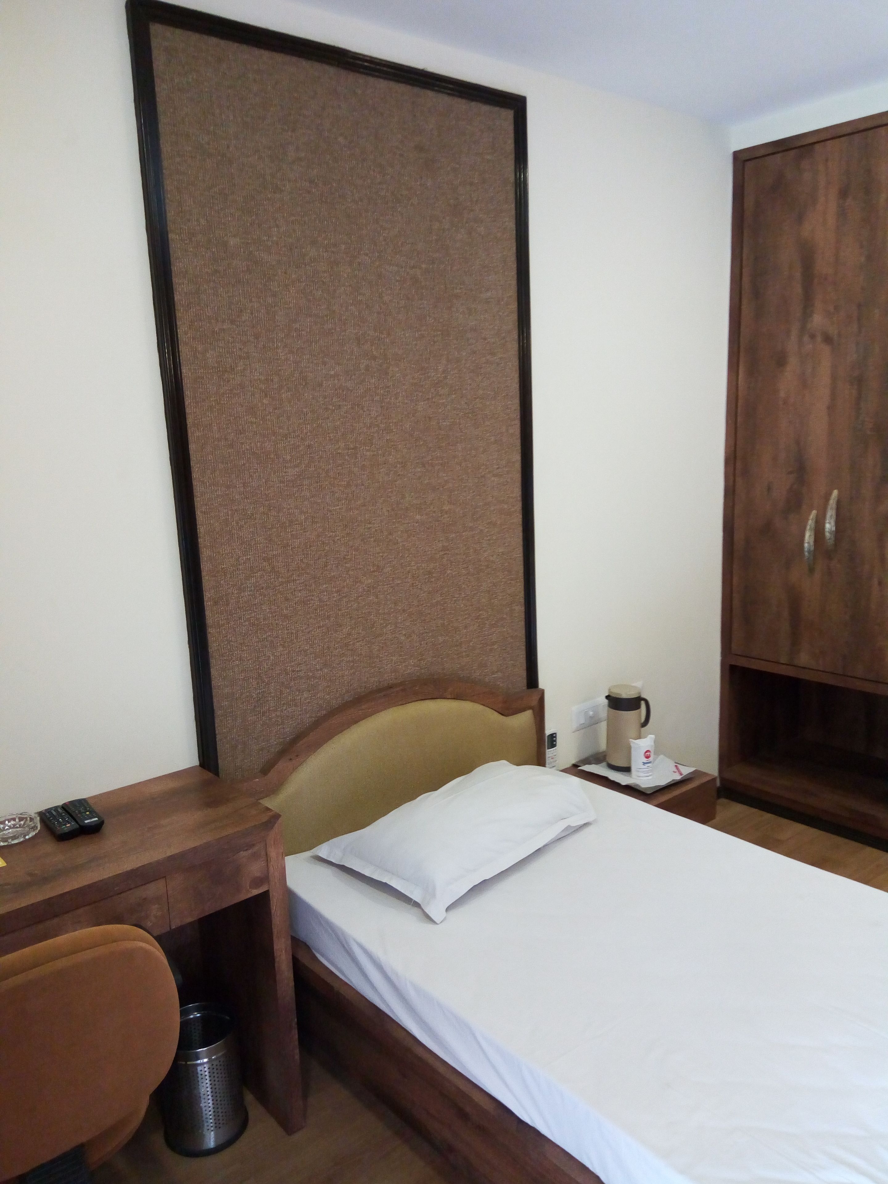 A hotel room with a wooden desk, a bed with a white pillow, a wooden wardrobe, and a small trash bin.