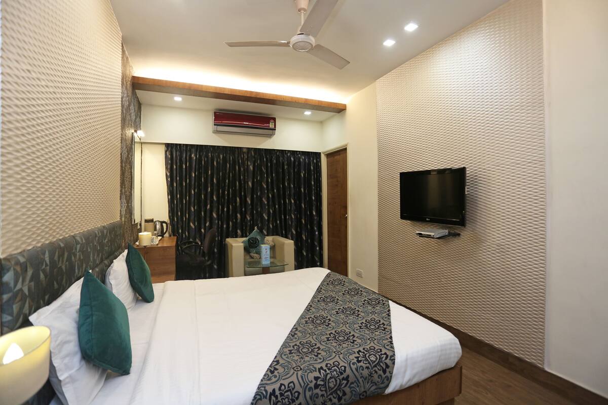 A hotel room with a bed, a flat-screen TV mounted on the wall, a ceiling fan, an air conditioning unit, and a small table with a chair.