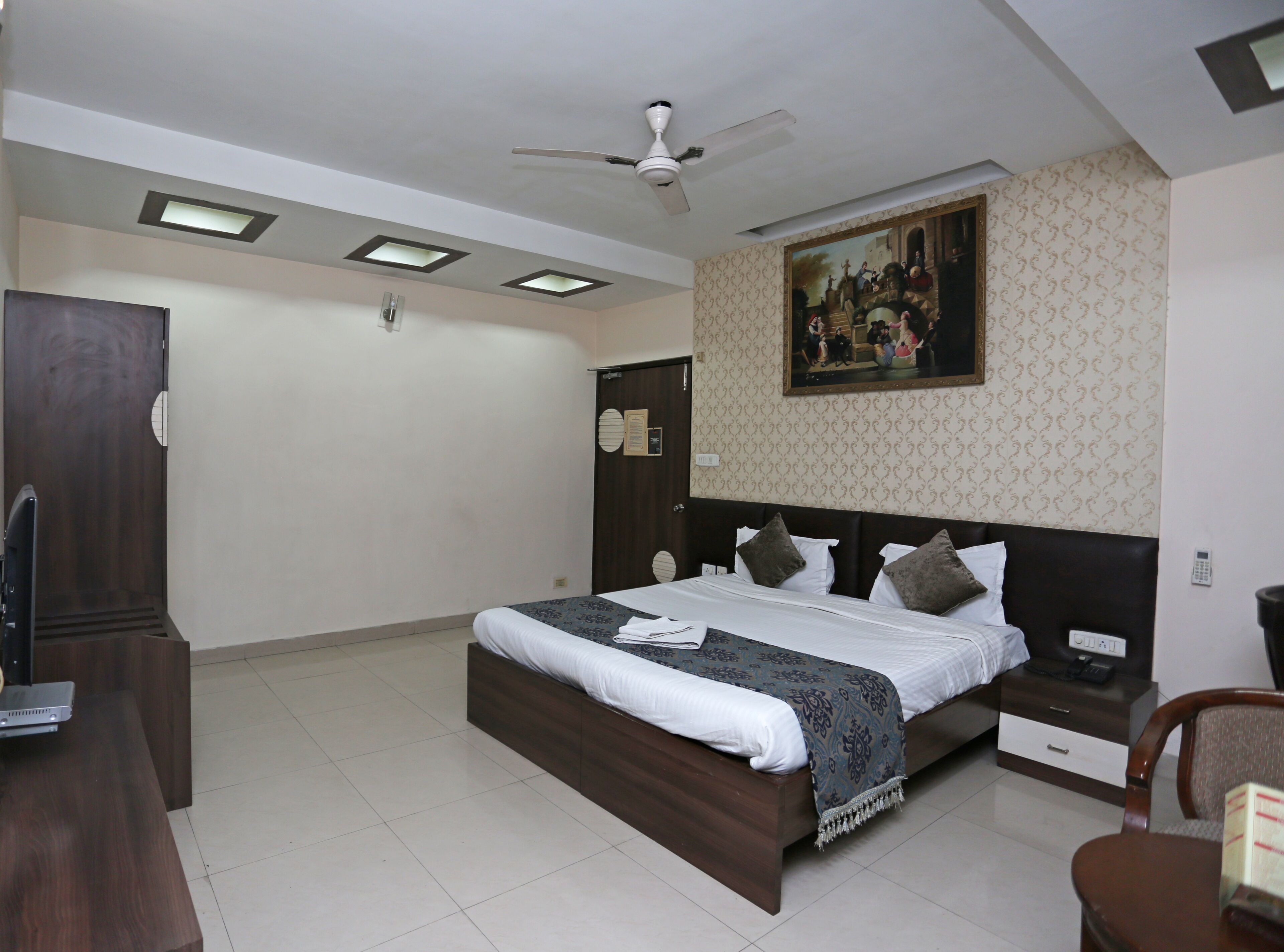 A bedroom with a bed, a nightstand, a desk, a chair, a ceiling fan, recessed lighting, and a wall-mounted painting.