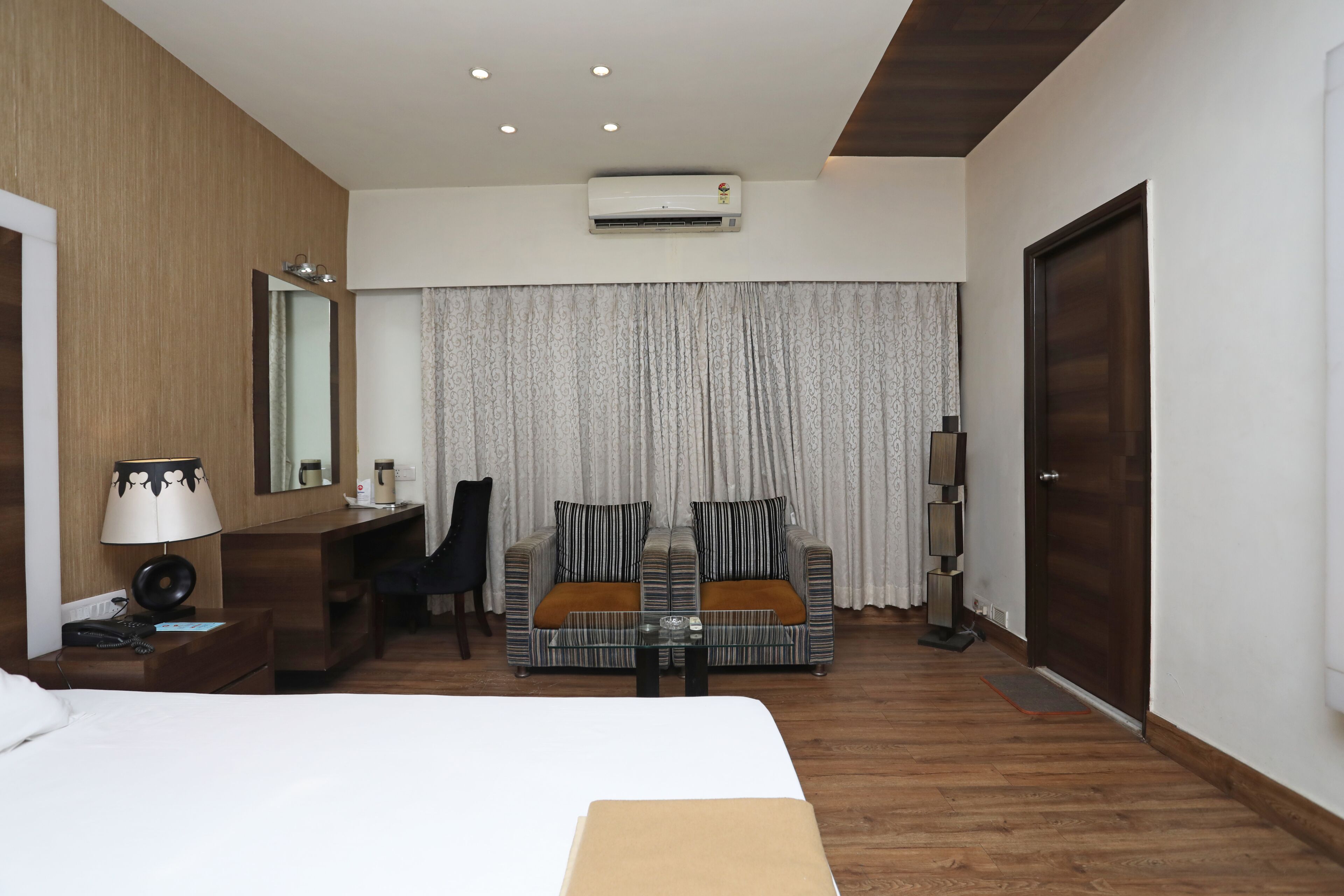 A hotel room with a bed, a desk, a sofa, a coffee table, a lamp, a mirror, an air conditioning unit, a door, and a curtain.