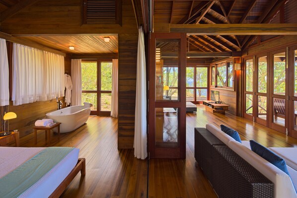 A spacious room with a large bed, a sitting area with a sofa, a bathtub, and large windows offering a view of the outdoors. - Secret Bay (Portsmouth)