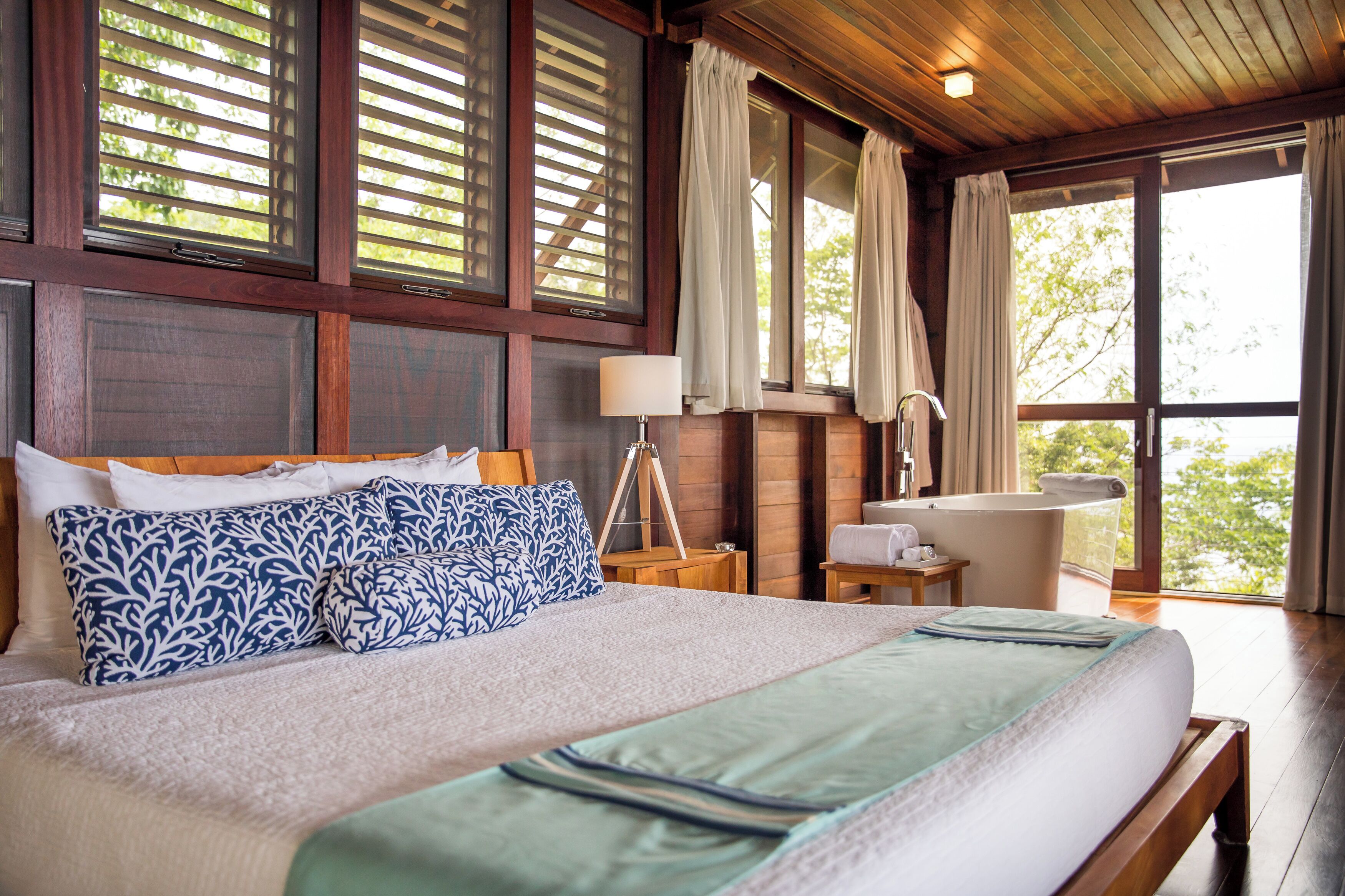 A bedroom with a large bed, a bedside table, a lamp, a window with curtains, and a view of the outdoors.