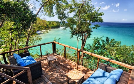 A wooden deck with wicker furniture and a view of a turquoise sea and lush greenery.