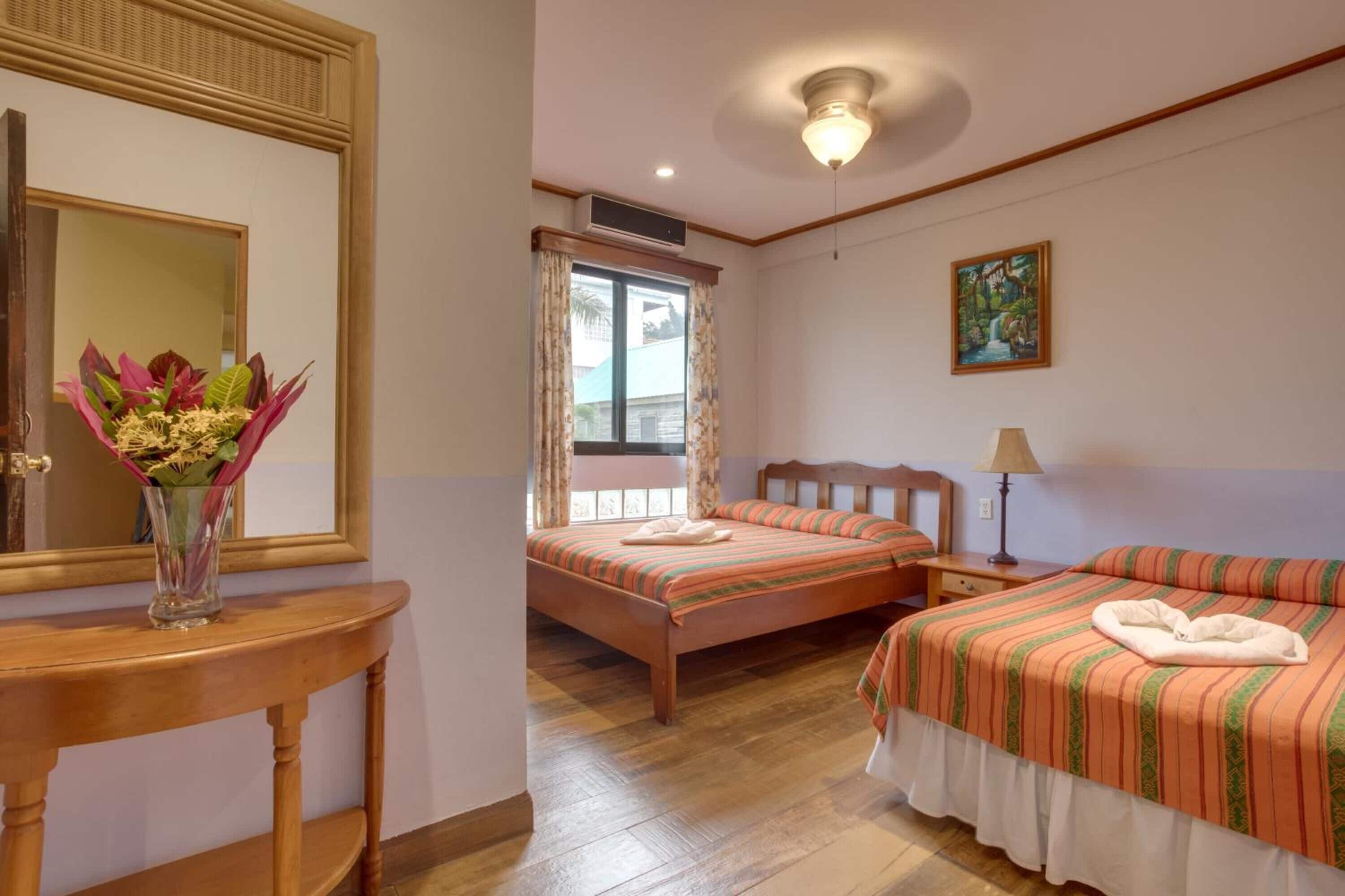 Una habitación de hotel con una cama de madera, una mesita de noche con lámpara, una ventana con cortinas, un espejo y un cuadro en la pared.