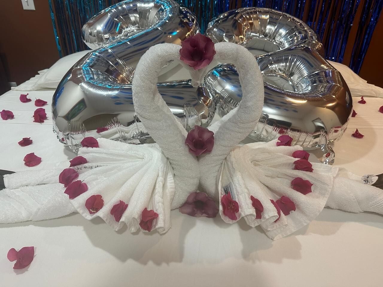 Una mesa con un centro decorativo que incluye toallas con forma de cisne y pétalos de rosa, rodeada de globos plateados.