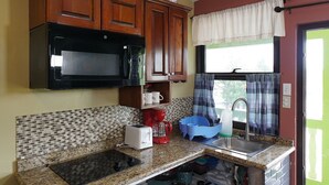 A kitchen with wooden cabinets, a black microwave, a red coffee maker, a white toaster, and a sink with a blue dish rack. - Venus Hotel (San Ignacio)