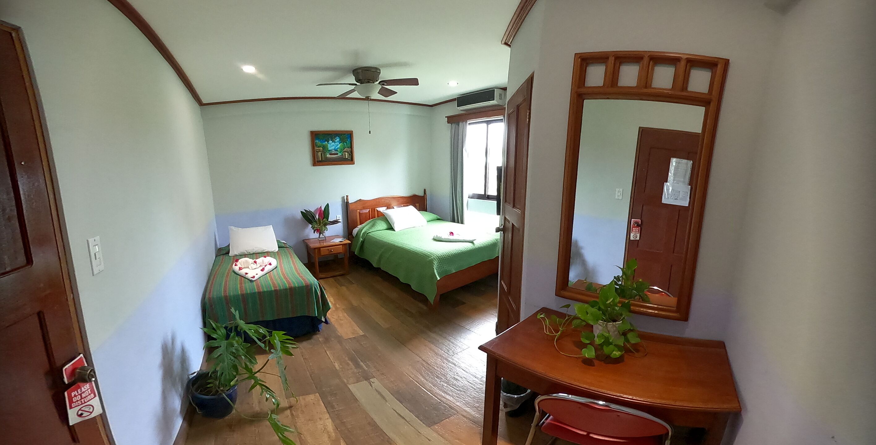 A room with two beds, a ceiling fan, a wooden desk, a mirror, and a potted plant.