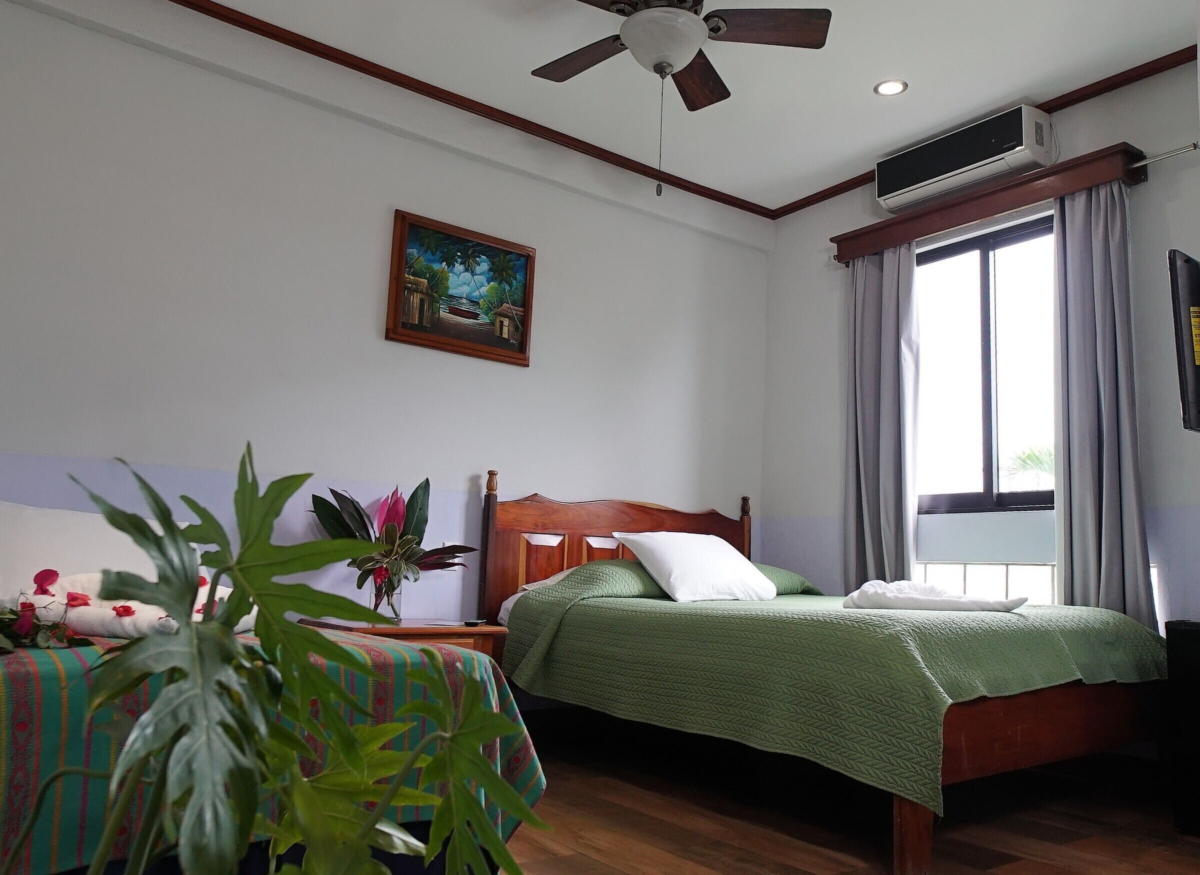 Een slaapkamer met een houten bed, een plafondventilator, een raam met gordijnen, een schilderij aan de muur en een nachtkastje met een plant.