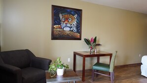 A living room with a dark sofa, a small table with a potted plant, a wooden chair, and a painting of a leopard on the wall. - Venus Hotel (San Ignacio)