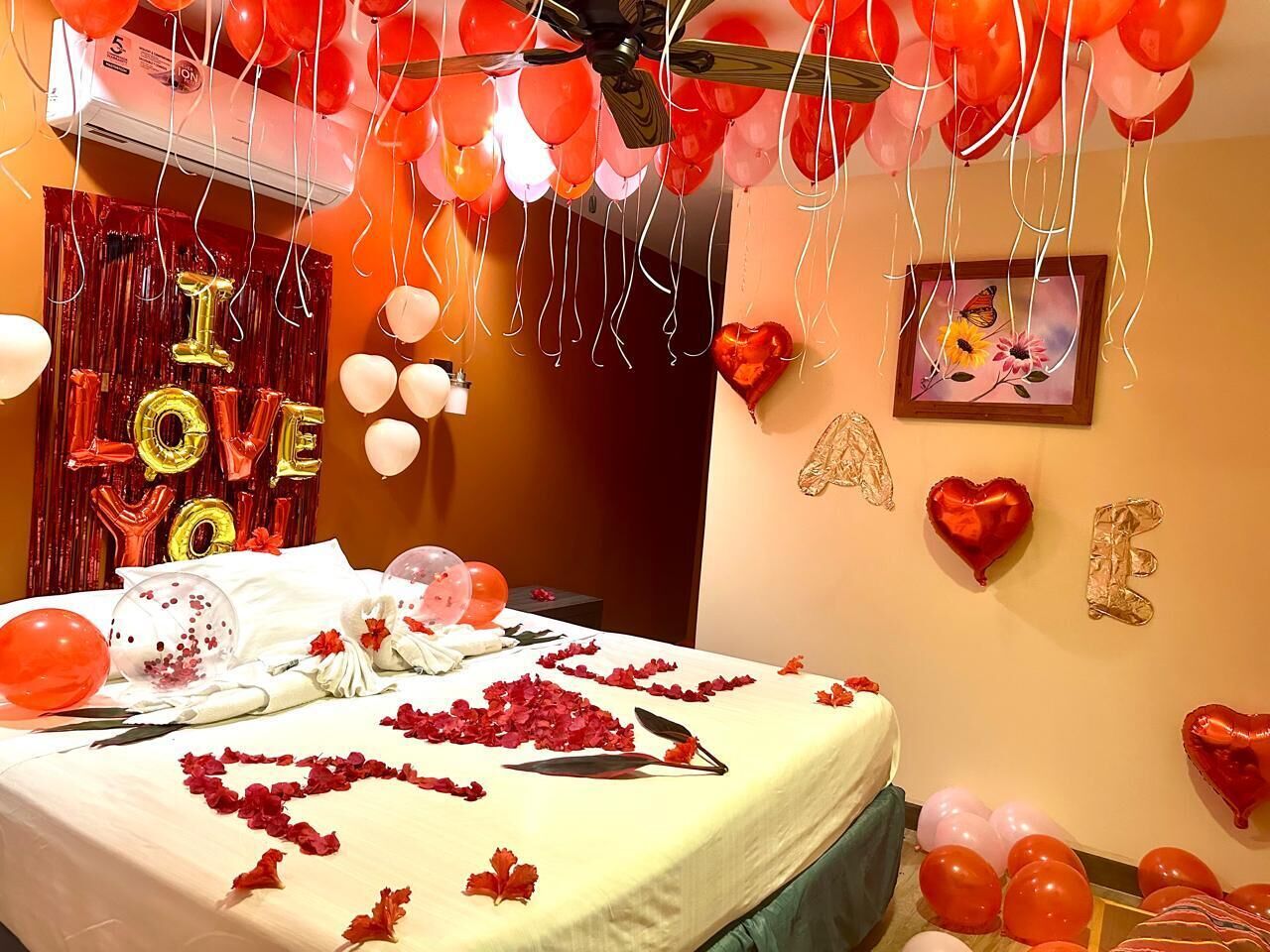 A room decorated with red and gold balloons, a bed with rose petals, and a 'I LOVE YOU' sign.