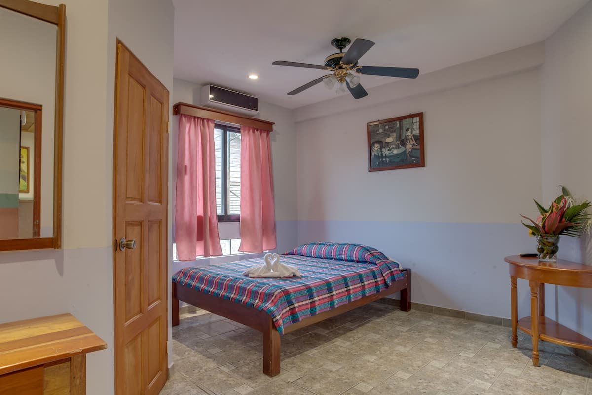 A bedroom with a bed, a ceiling fan, a window with curtains, a bedside table, and a wall-mounted air conditioner.