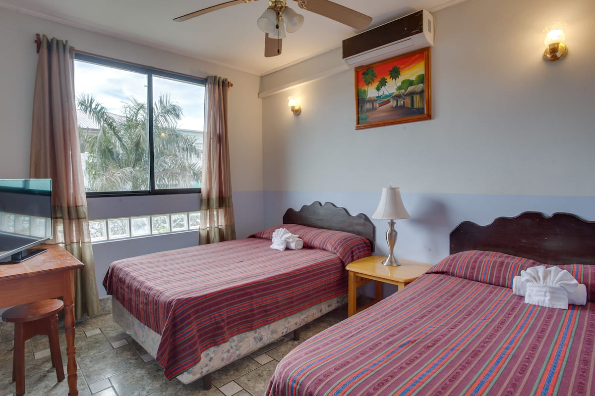 A hotel room with two beds, a desk, a ceiling fan, a window with curtains, a painting on the wall, and a lamp on the nightstand.