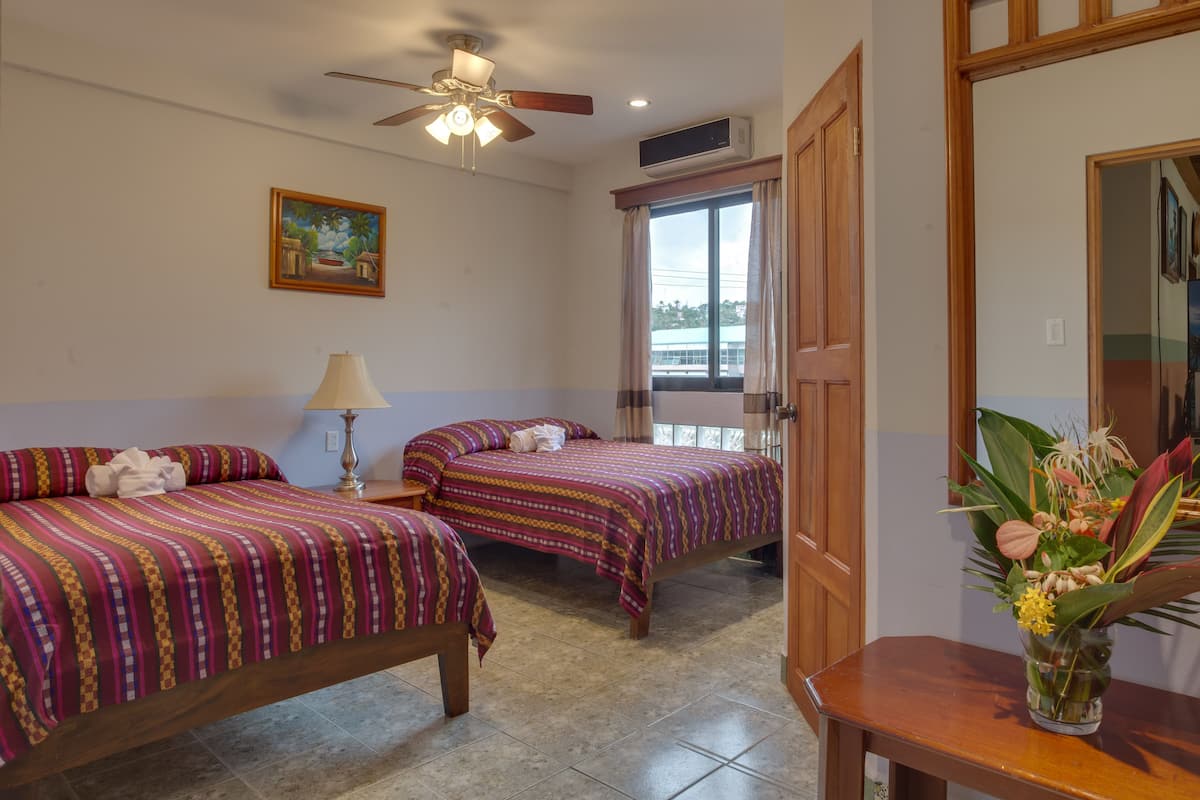 A hotel room with two beds, a ceiling fan, a window with curtains, a wooden door, and a vase with flowers on a table.