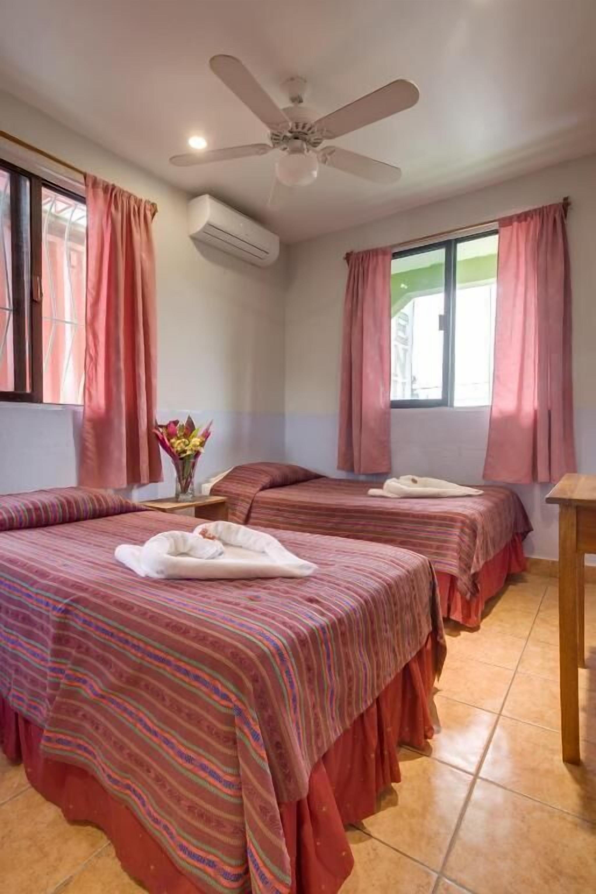 A hotel room with two beds, pink curtains, a ceiling fan, and an air conditioning unit.