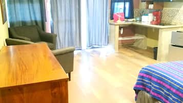 A compact room with a wooden desk, a chair, a bed with a striped blanket, a kitchenette with a microwave, and a window with curtains.