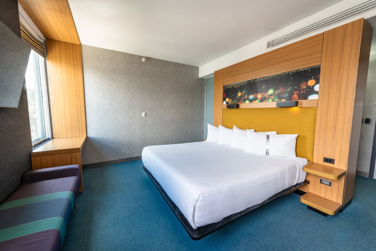 A modern hotel room with a large bed, a small sofa, a wooden headboard, and a window with blinds.