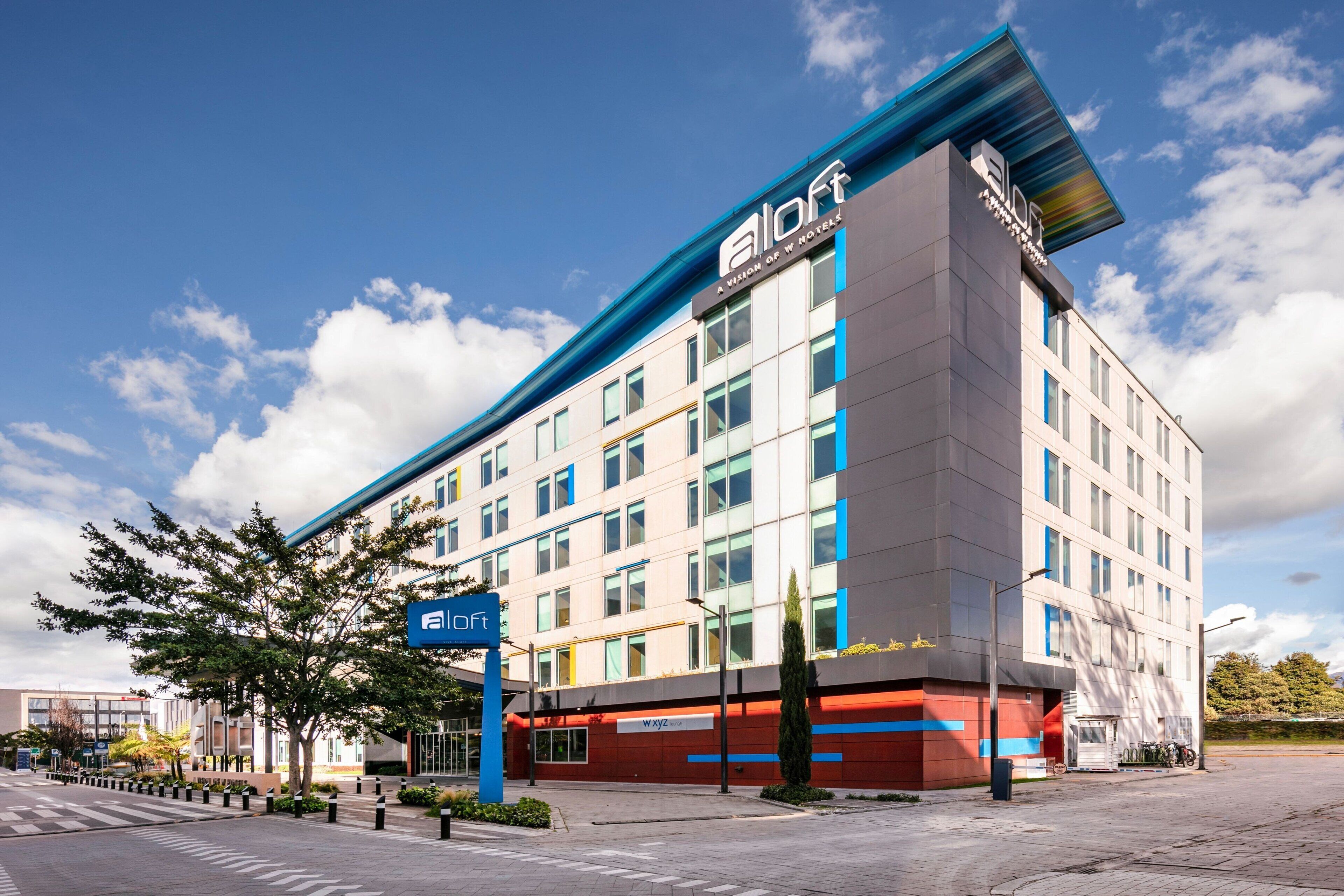 A modern hotel building with a prominent 'aloft' sign, multiple floors, and a clear sky.